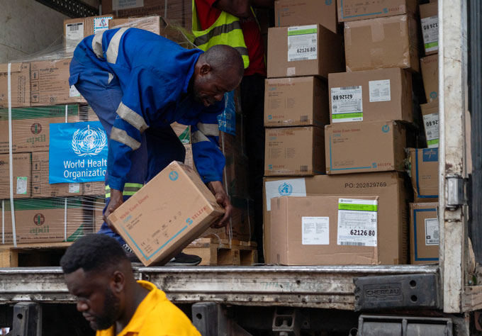 A worker unloads cardboard boxes of medical supplies from a delivery truck in the Democratic Republic of Congo, while another worker walks in the foreground.